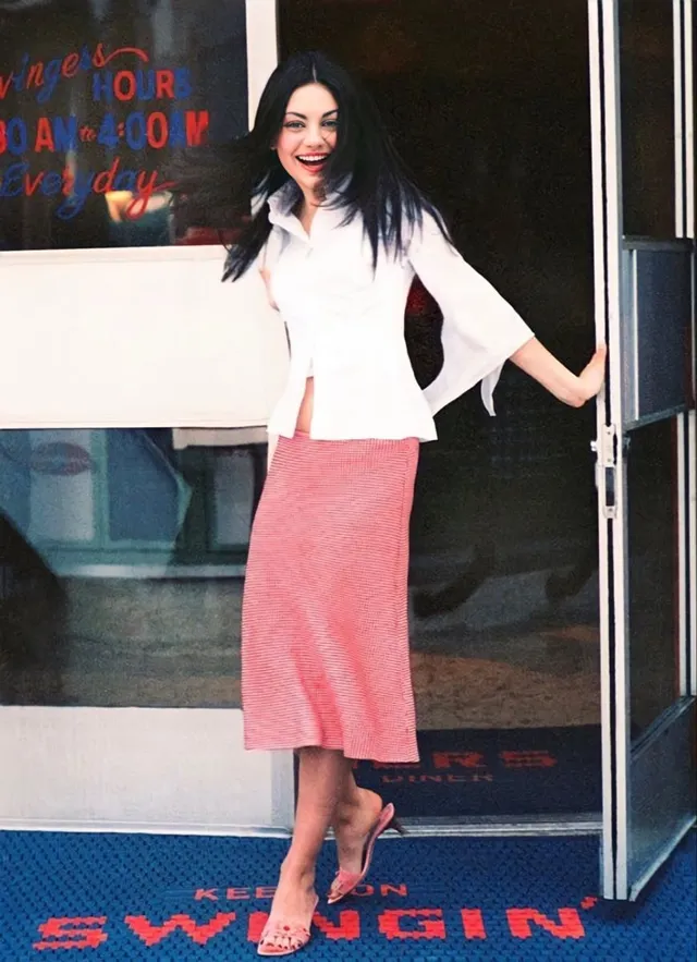Mila Kunis posing at an event in a white blouse, pink skirt and heels.