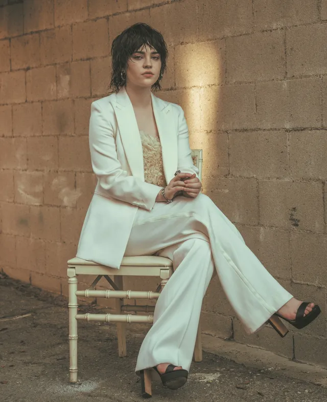 Sophie Thatcher sitting in a white trouser suit and platform heels.