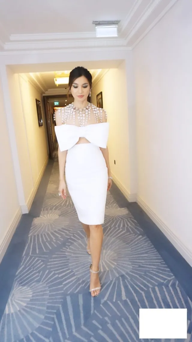 Gemma Chan standing in a hallway in a white dress.