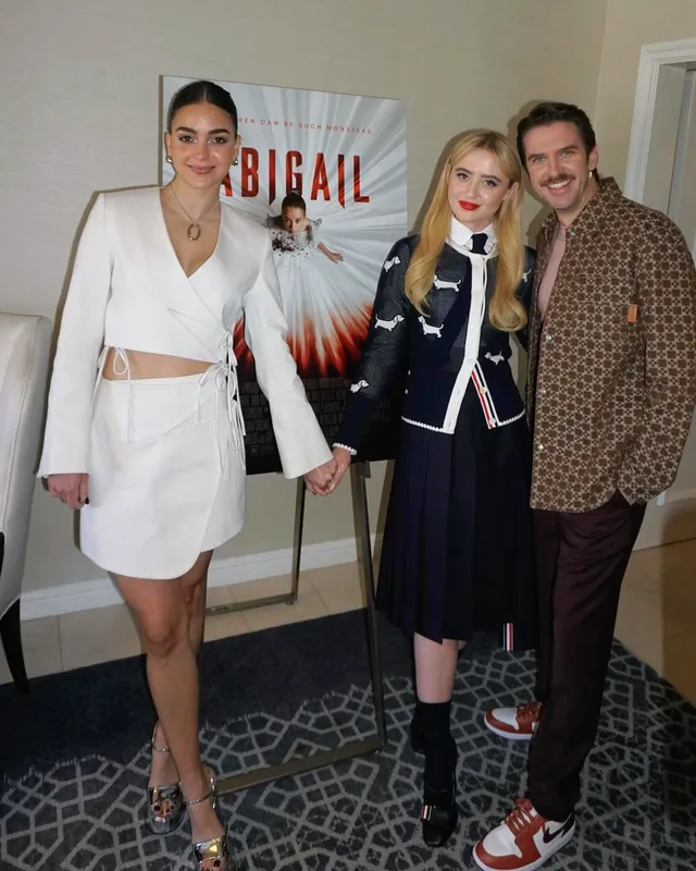 Melissa Barrera posing with two people in a white outfit and metallic heels.