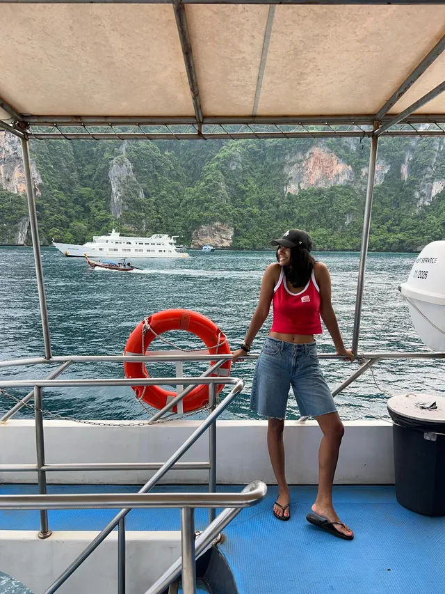 Greta Onieogou standing on a boat in a red top, denim shorts and flat sandals.
