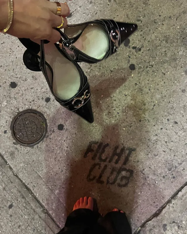 Brec Bassinger shown standing barefoot beside a pair of black heels on a pavement.