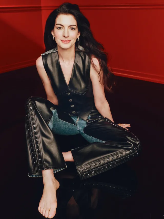 Anne Hathaway sitting barefoot in a black outfit on a chair.