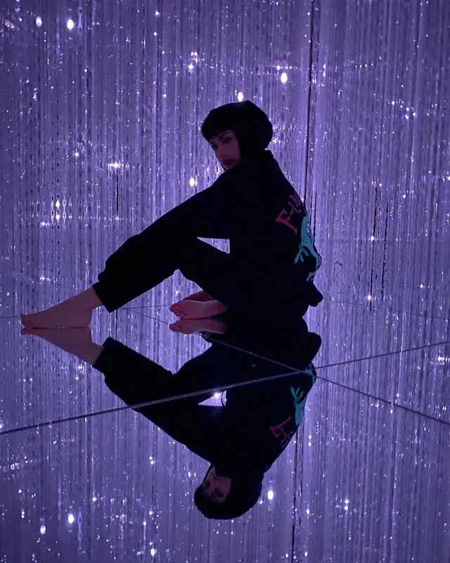 Úrsula Corberó sitting barefoot on a reflective surface against a purple backdrop.