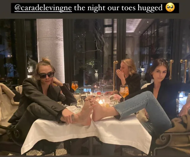 Cara Delevingne sitting at a restaurant with a companion and their feet raised.