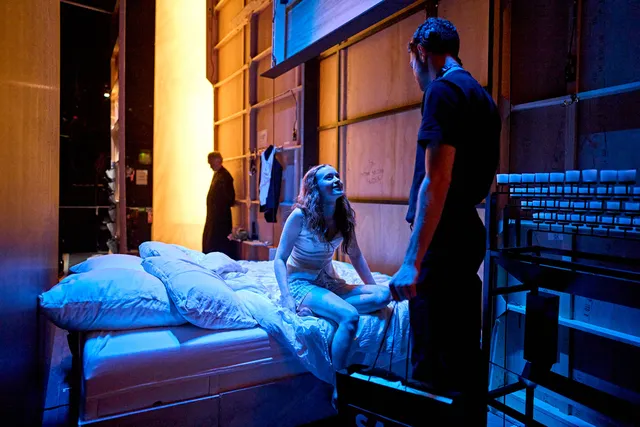 Sadie Sink sitting barefoot on a bed in a blue lit stage scene.