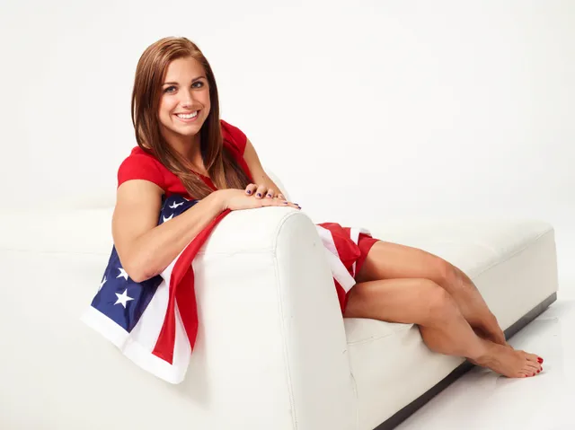 Alex Morgan sitting barefoot on a studio sofa holding a football.