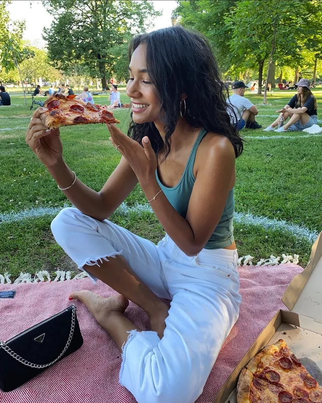 Greta Onieogou sitting barefoot on a picnic blanket while eating pizza.