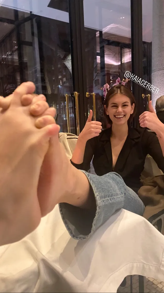 Cara Delevingne sitting at a table with one foot toward the camera.