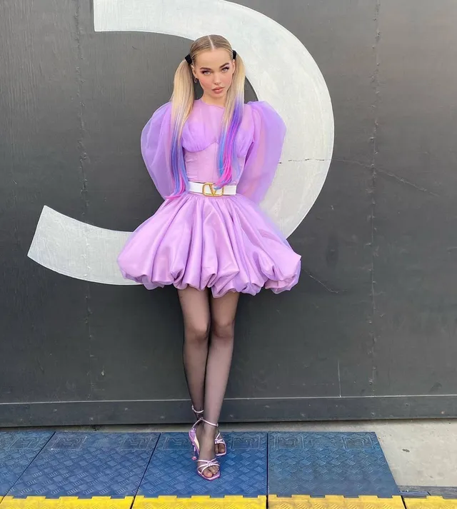 Dove Cameron standing in a purple dress and clear heels outside.