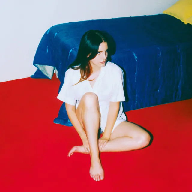 Lana Del Rey sitting barefoot beside a bed with a red floor.