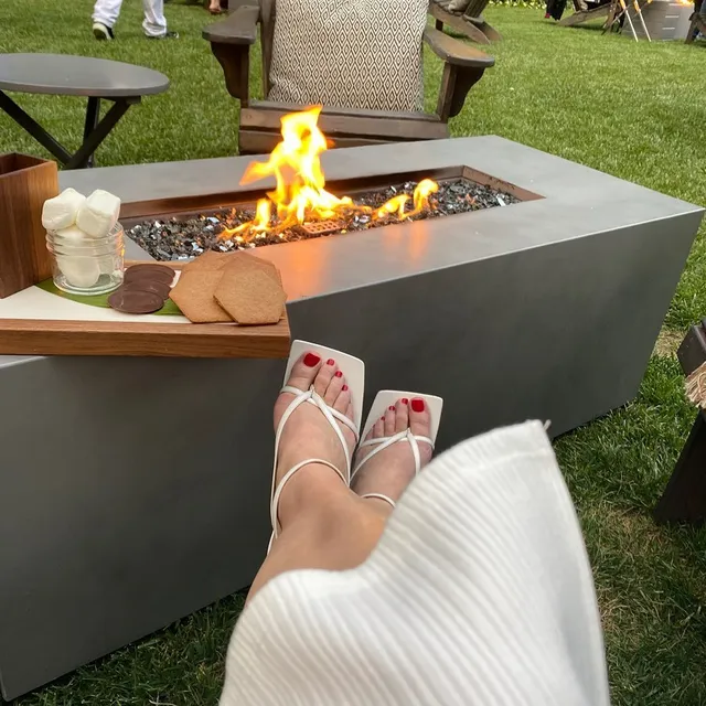 Halston Sage shown wearing white strappy flat sandals beside a fire pit.