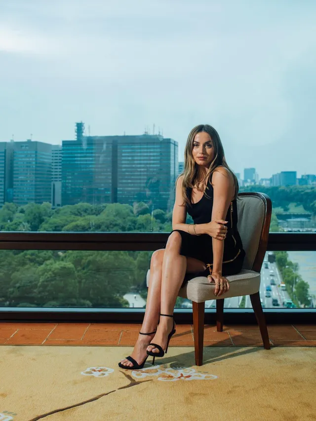 Ana de Armas seated by a city-view window in a black dress and heeled sandals.