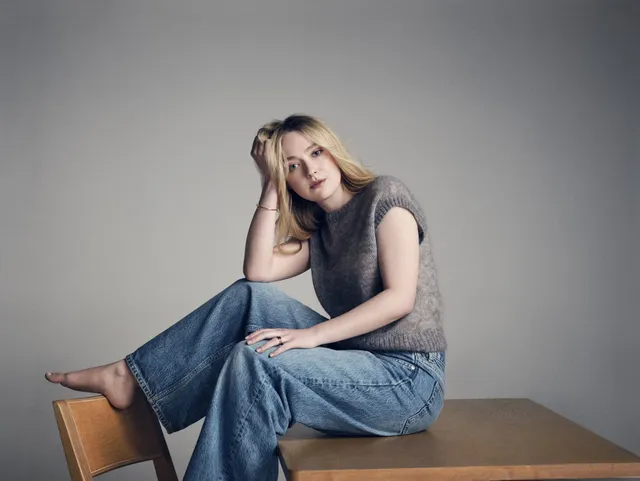 Dakota Fanning sitting barefoot on a table in a gray top and jeans.