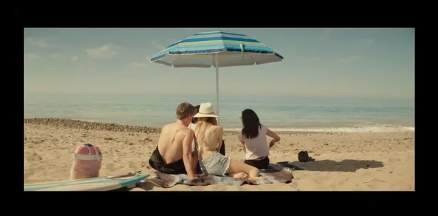 Halston Sage sitting barefoot with a group on a beach in a scene.