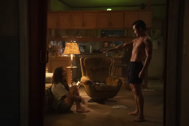 Adria Arjona sitting beside a bed in a dimly lit film scene.