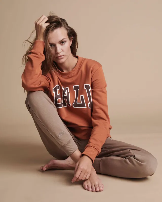 Josephine Skriver sitting barefoot in a studio wearing an orange sweatshirt.