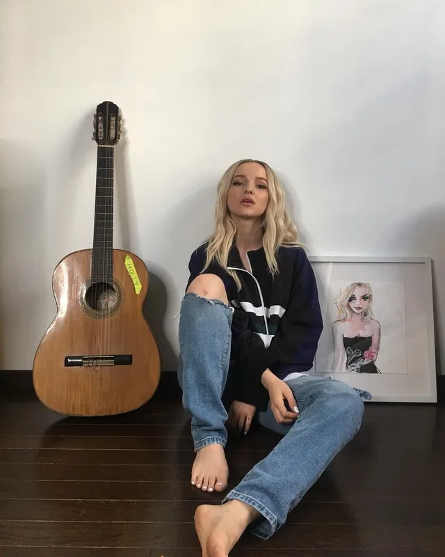Dove Cameron sitting barefoot beside a guitar in jeans.