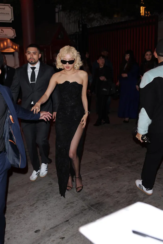 Rosé walking through a nighttime crowd in a strapless black gown and heeled sandals.