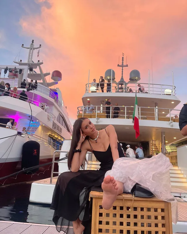 Nina Dobrev sitting barefoot on a boat deck at sunset.
