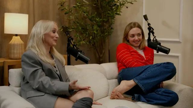 Amanda Seyfried sitting cross-legged barefoot on a couch beside another guest.