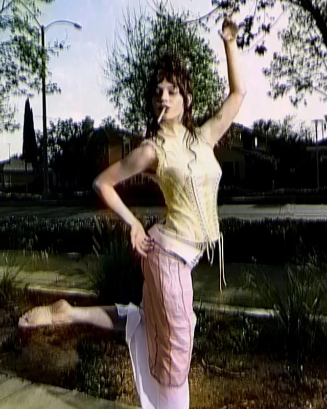 Sophie Thatcher posing outdoors in pink trousers with one foot raised.