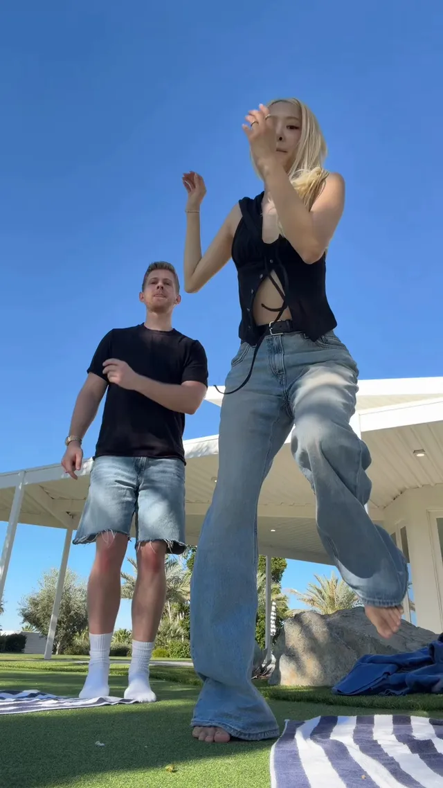 Rosé dancing barefoot on a lawn beside another person.