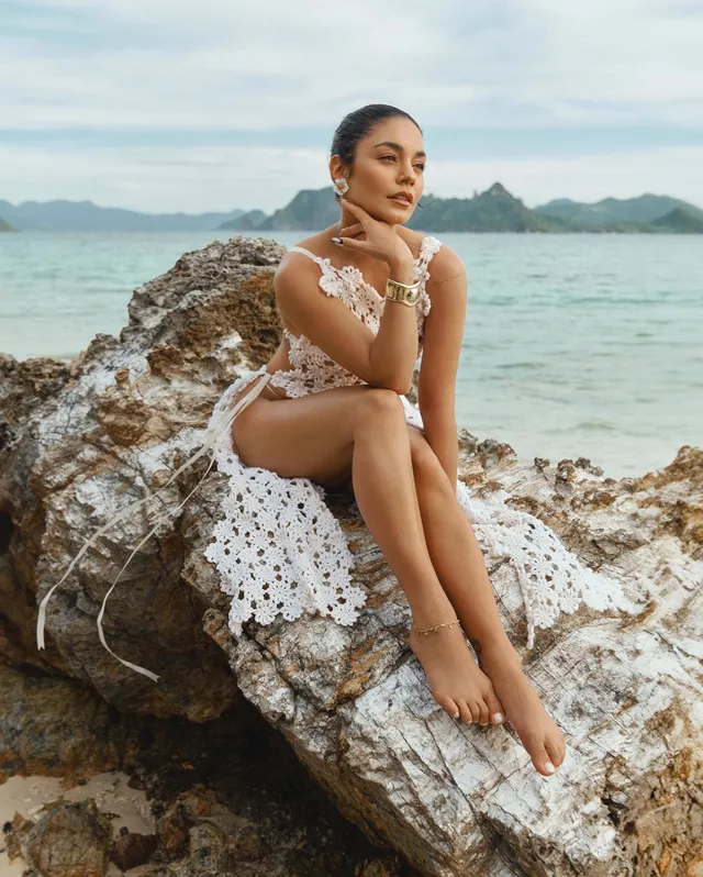 Vanessa Hudgens standing barefoot on coastal rocks in a white flowered dress.
