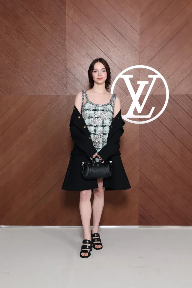 Emma Myers standing beside a wooden event wall in black platform heels.