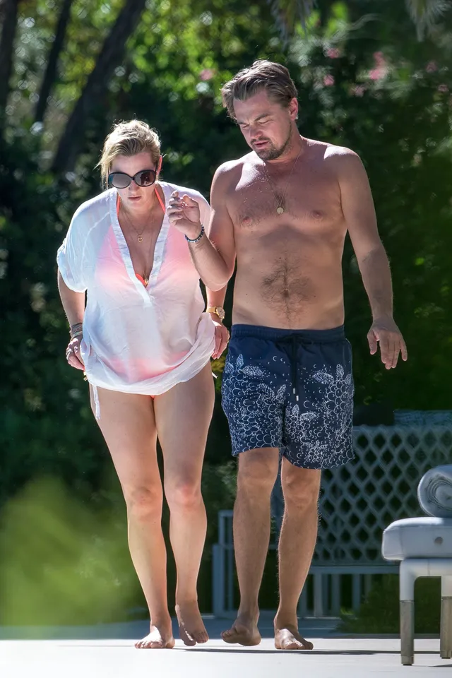 Kate Winslet walking barefoot outdoors in a white cover-up beside a companion.