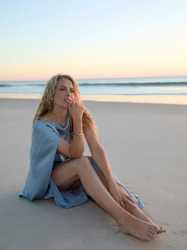 Teresa Palmer sitting on a sandy beach in a blue dress.