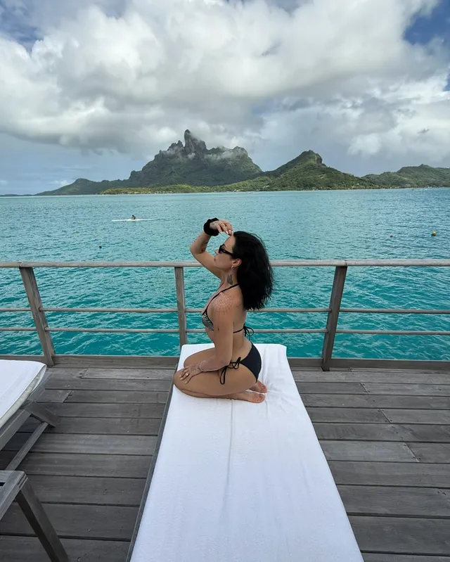 Demi Lovato kneeling barefoot on a lounger above turquoise water.