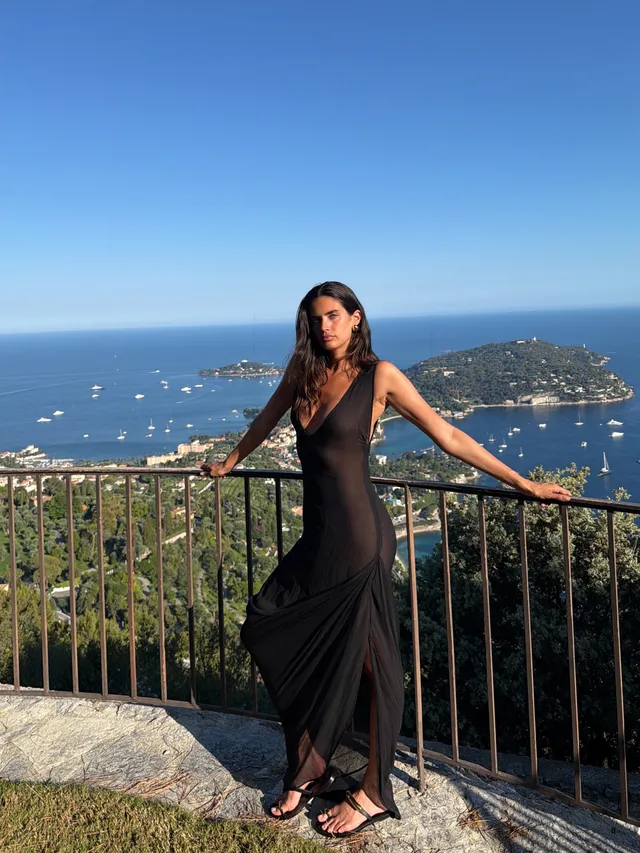 Sara Sampaio standing in flat sandals beside a terrace railing wearing a black dress.