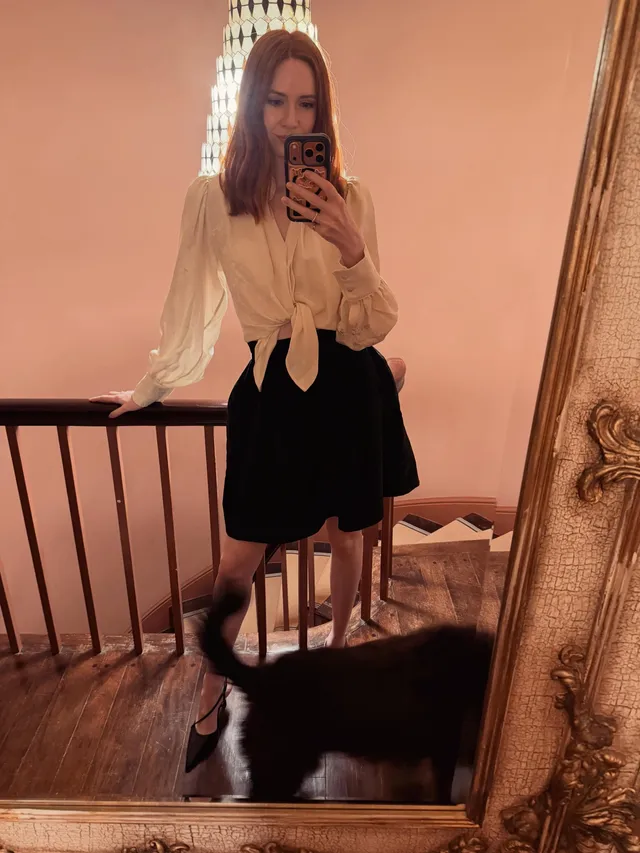 Karen Gillan taking a mirror photo in a white blouse and black heels.