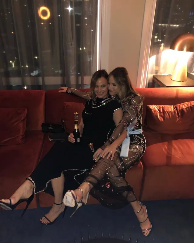 Halston Sage sitting beside a friend on an orange sofa in a patterned dress and heels.