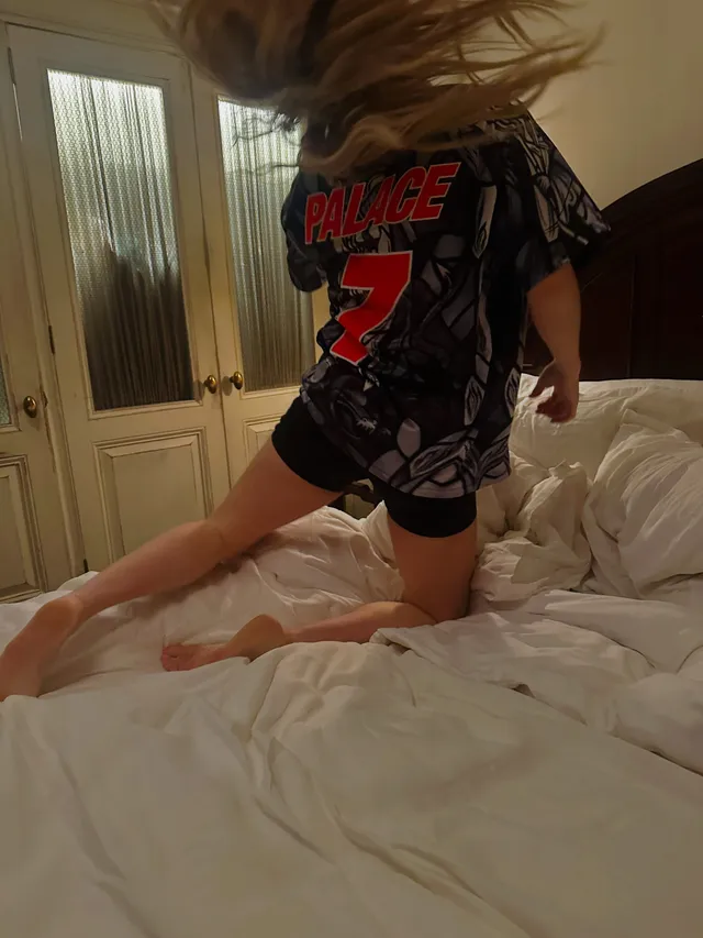Ellie Goulding jumping barefoot on a bed in a dark shirt and shorts.