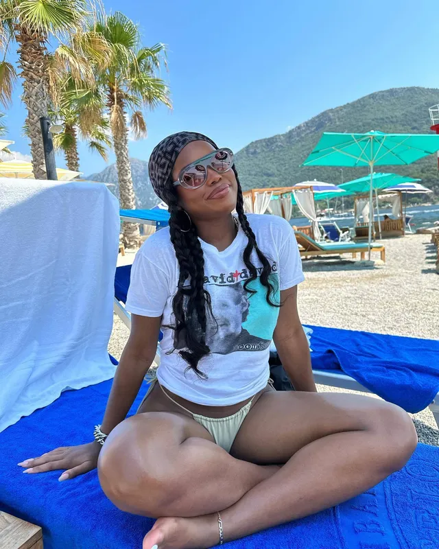 Ryan Destiny sitting barefoot on a beach lounger in a T-shirt and bikini bottoms.