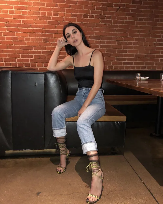 Adelaide Kane sitting on a bench in jeans and gold sandals.