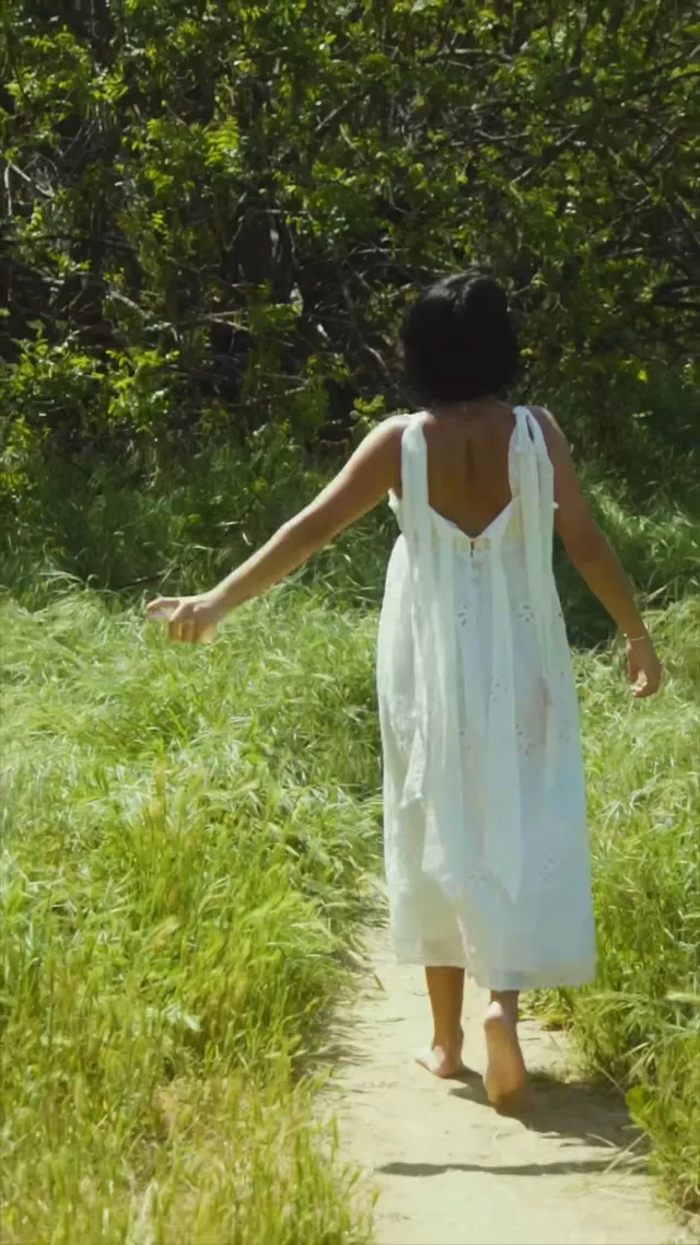 Isabela Merced walking barefoot along a garden path in a pale green dress.