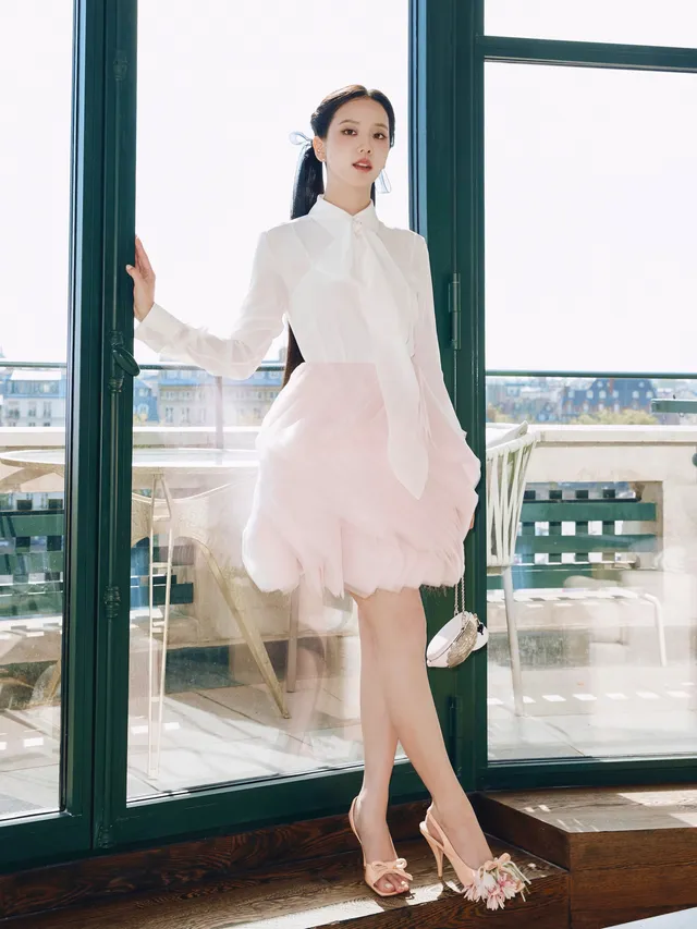 Jisoo wearing pale embellished heels and a pink skirt beside a balcony door.