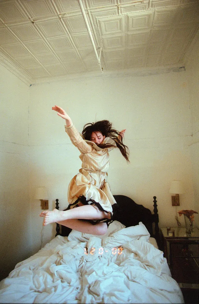Sophie Thatcher jumping barefoot on a bed in a pale dress.