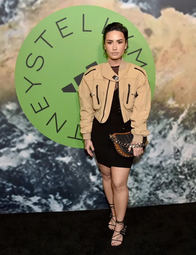Demi Lovato standing at an event in a tan jacket, short skirt and heels.