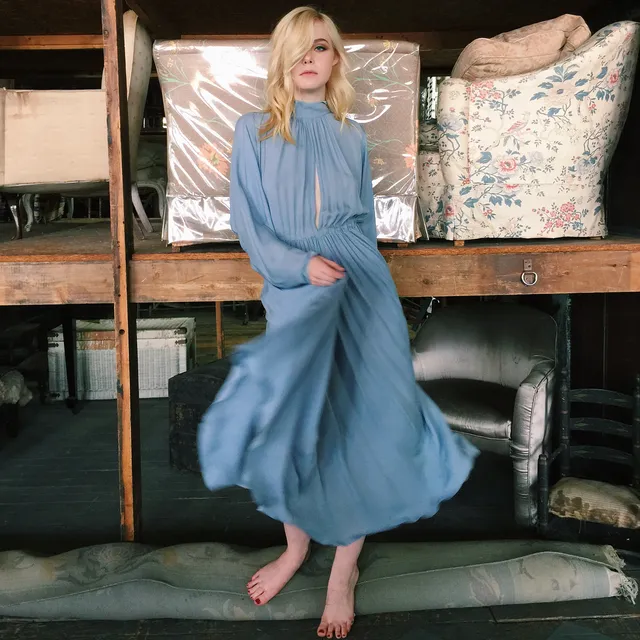 Elle Fanning sitting barefoot in a blue dress indoors.