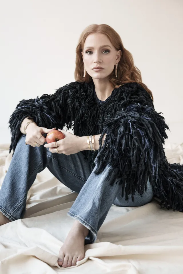 Jessica Chastain sitting on a bed in jeans and a feathered jacket.