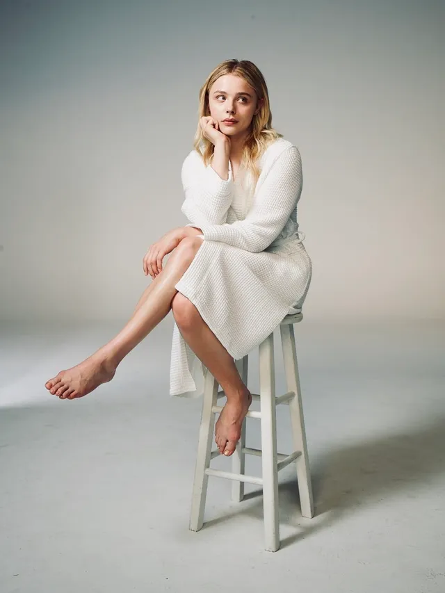 Chloë Grace Moretz sitting barefoot on a stool in a white dress.
