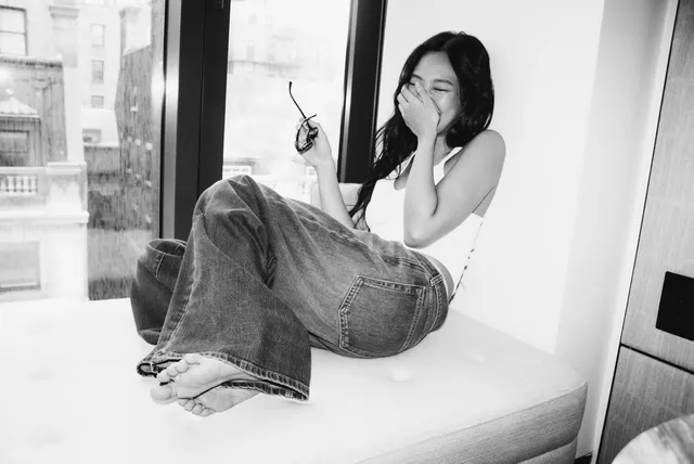 Jennie Kim sitting barefoot on a window seat in a monochrome photograph.