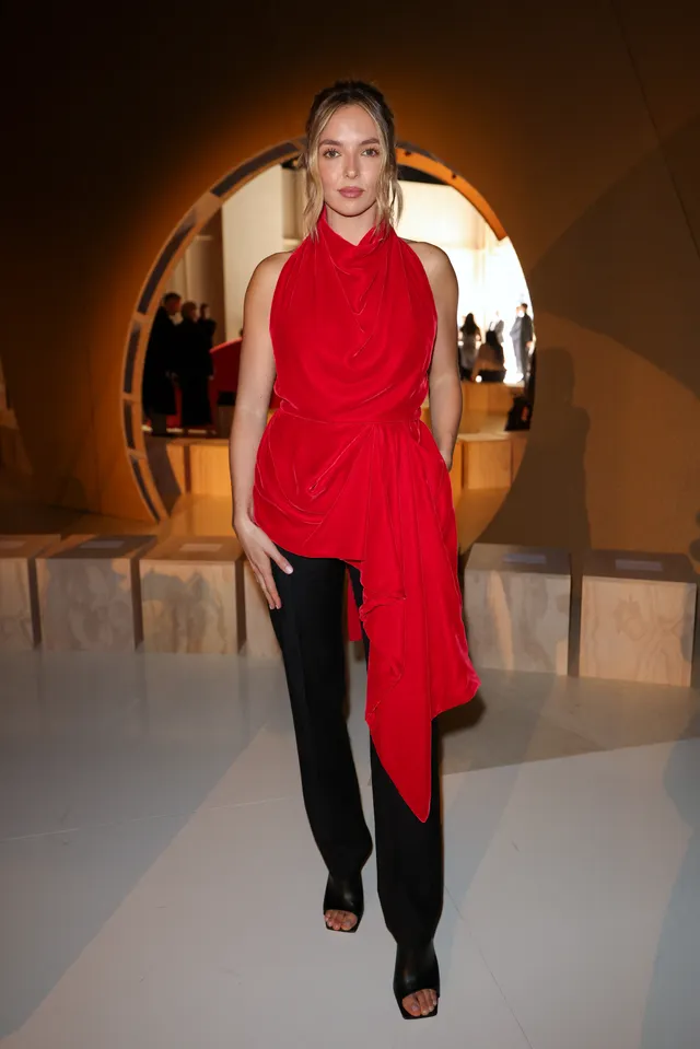 Jodie Comer posing indoors in a red top and black trousers.