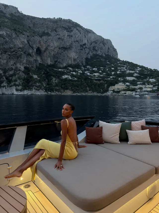 Jasmine Tookes reclining barefoot on a terrace seat in a yellow dress.