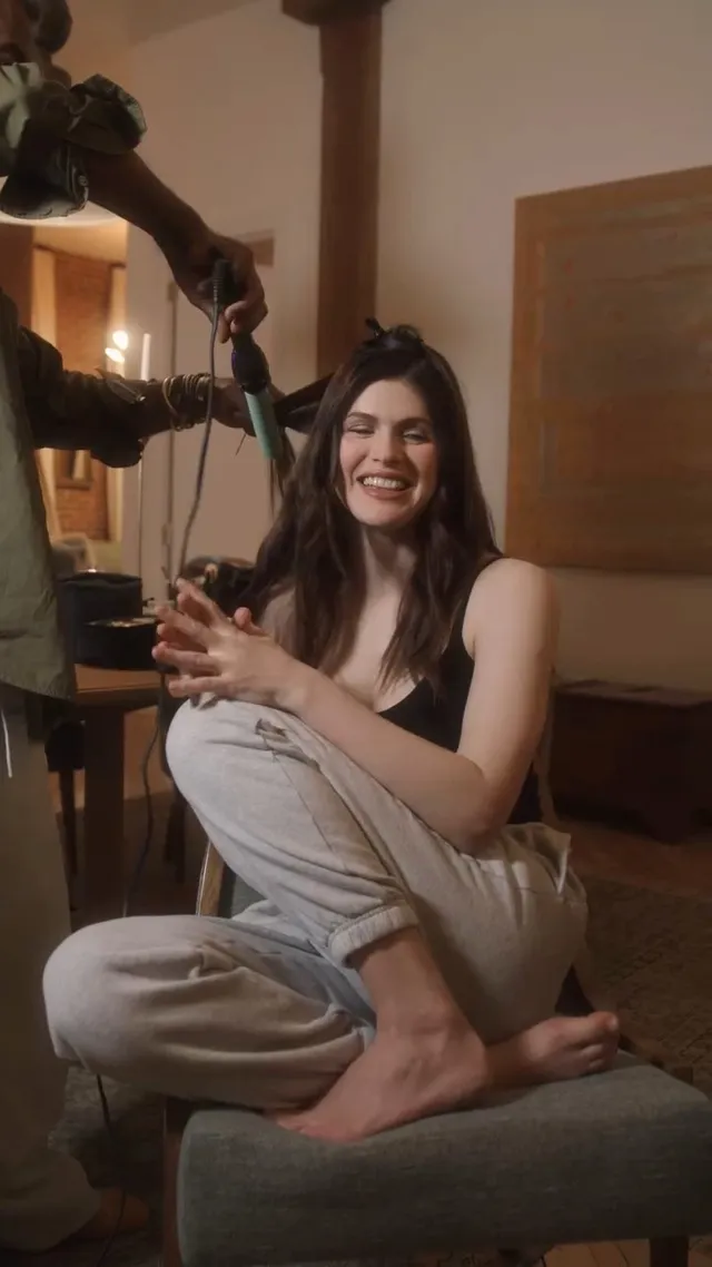 Alexandra Daddario sitting barefoot in a chair in a gray outfit.