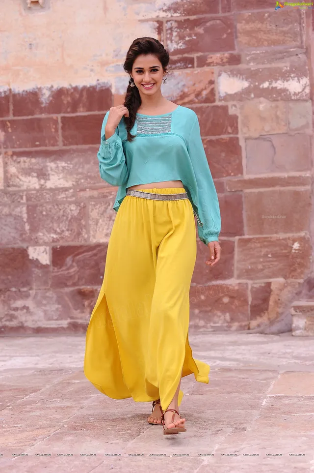 Disha Patani walking in flat sandals with yellow trousers and a turquoise blouse.